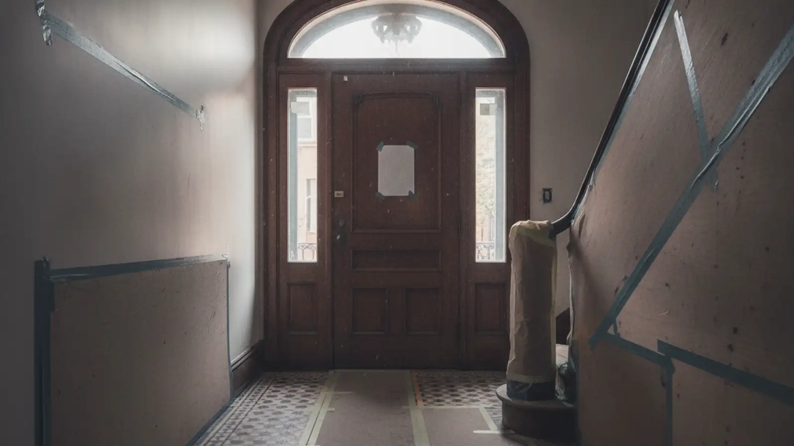 The vestibule of a brownstone with its original door and tile floor covered in protective board, daylight through the transom.